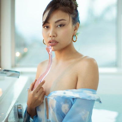 Young Asian woman in bathtub holding curved borosilicate glass dildo pink pressed against her lips